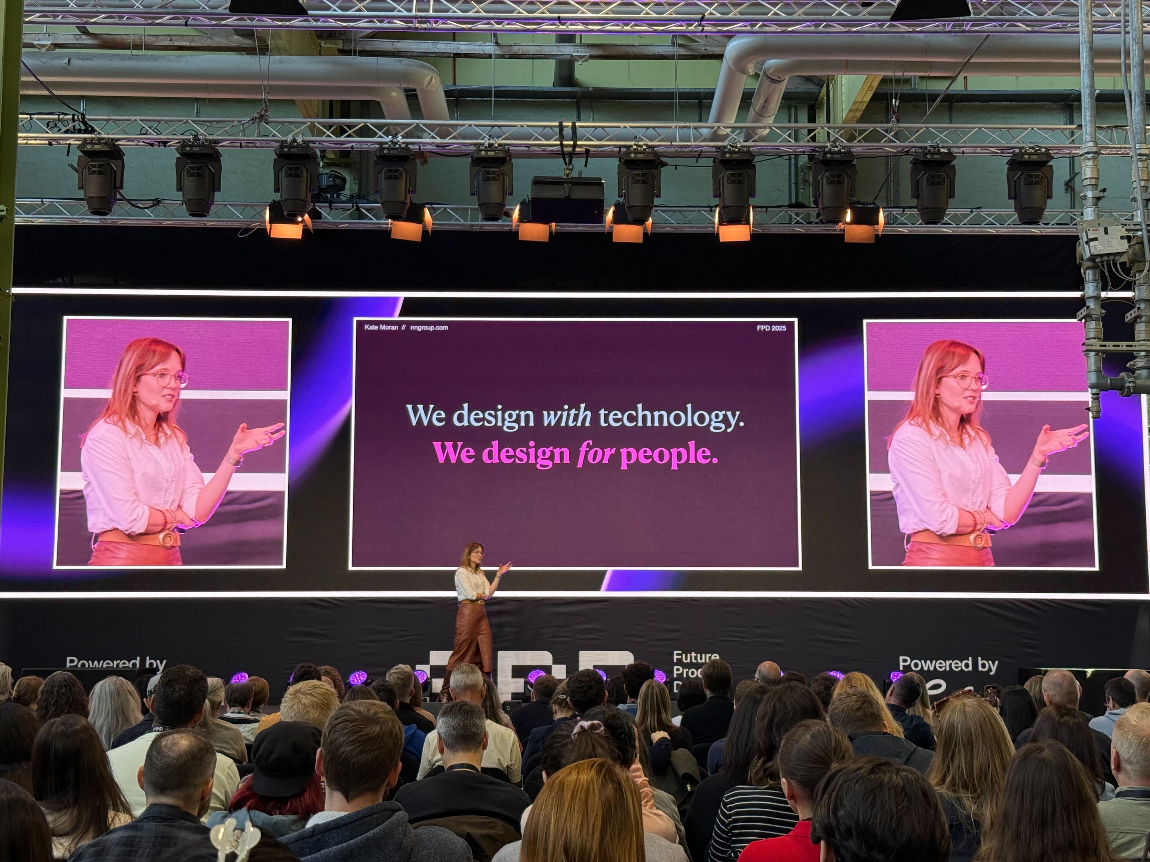 Conference presenter on stage with purple screens showing 'We design with technology for people'