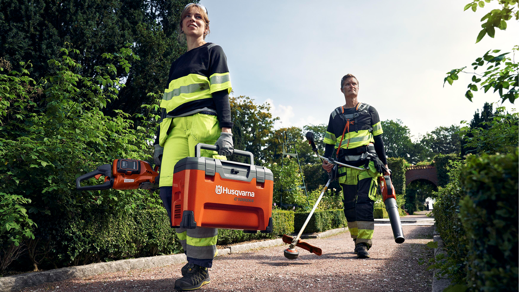 Two workers in hi-vis gear carrying Husqvarna equipment on tree-lined path
