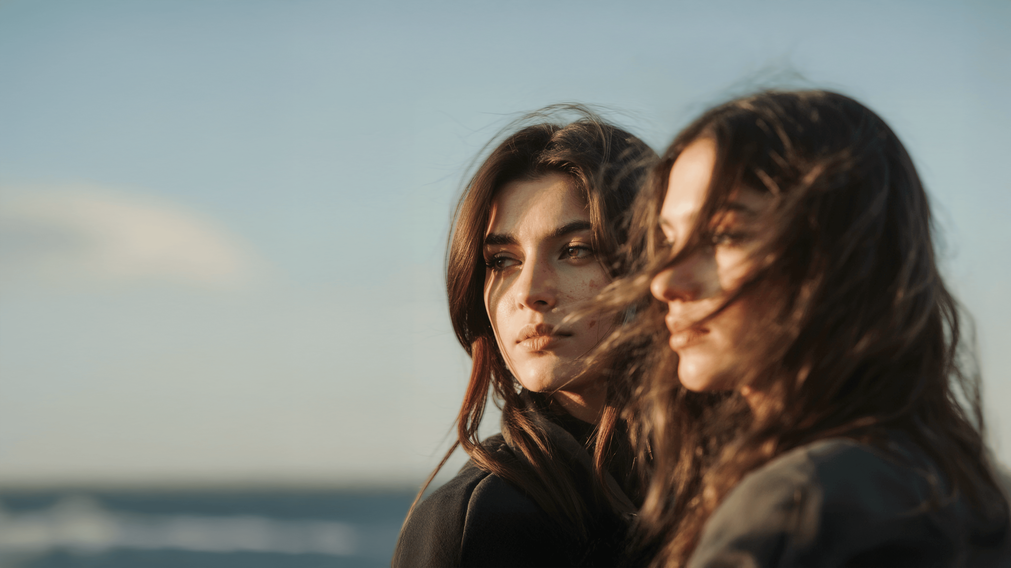 Two women with long hair stand close together outdoors, gazing in the same direction.