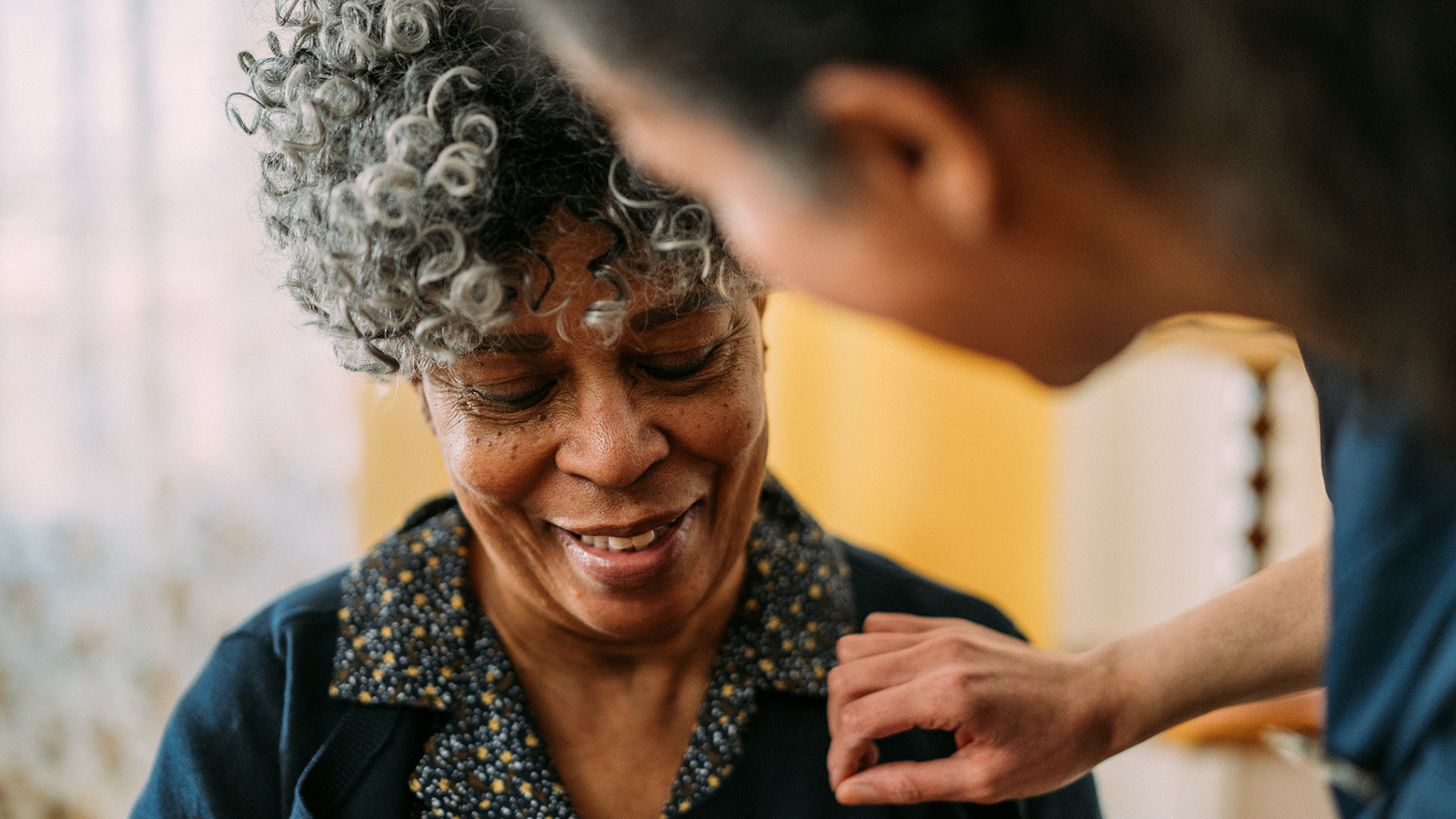 Smiling elderly woman with curly gray hair receiving gentle care from healthcare worker
