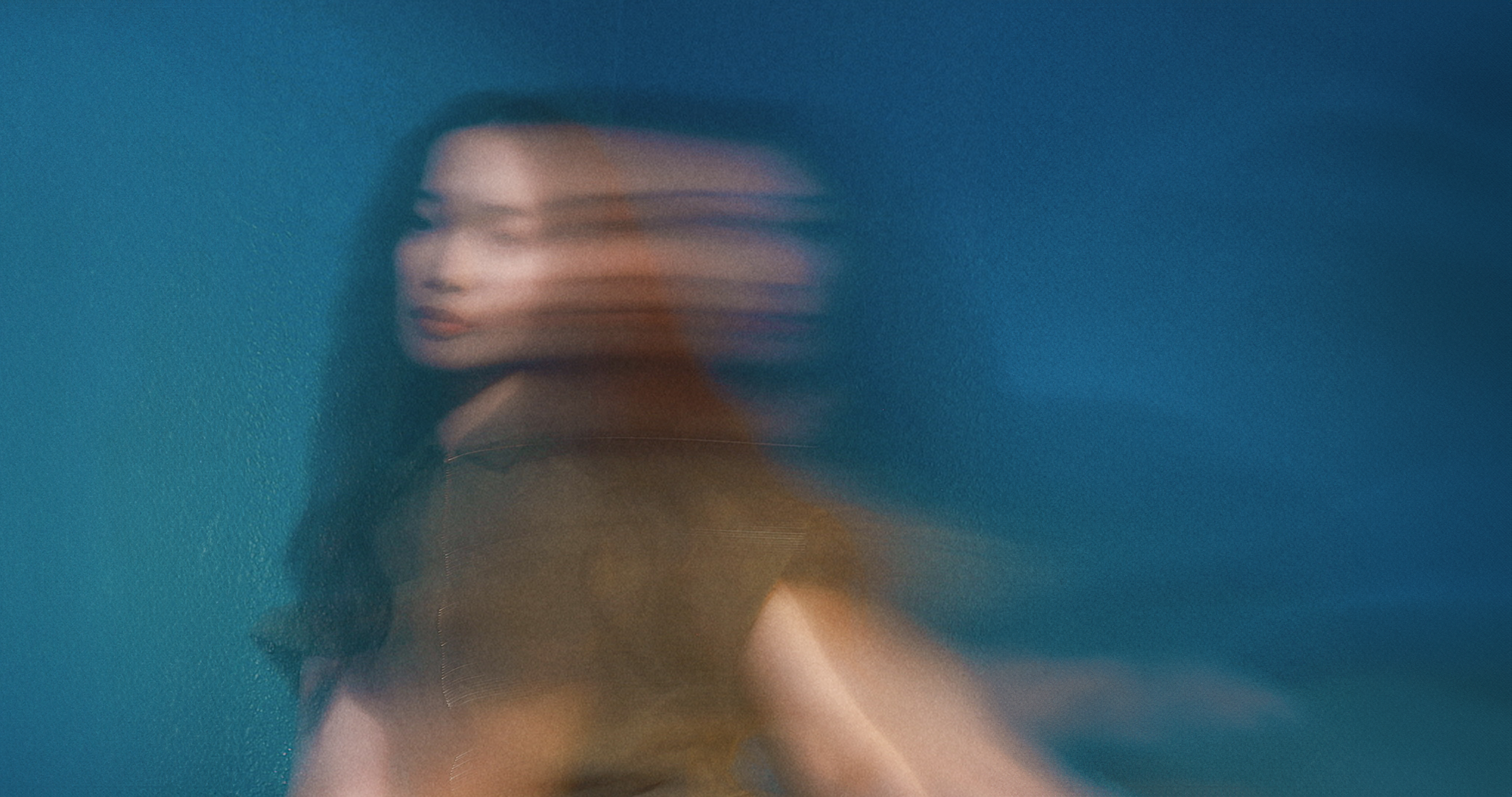 Figure in brown clothing with intentional motion blur against deep blue background, creating an abstract, ethereal effect.