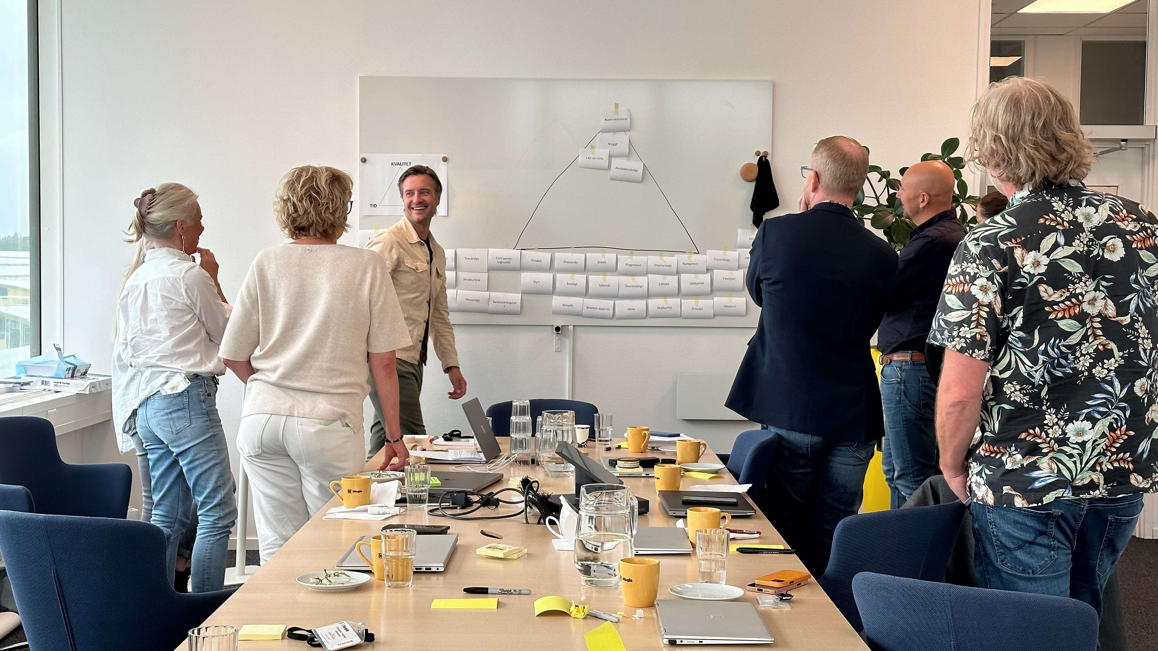 People participate in a brainstorming session around a table, with laptops and sticky notes. A wall shows organized papers.