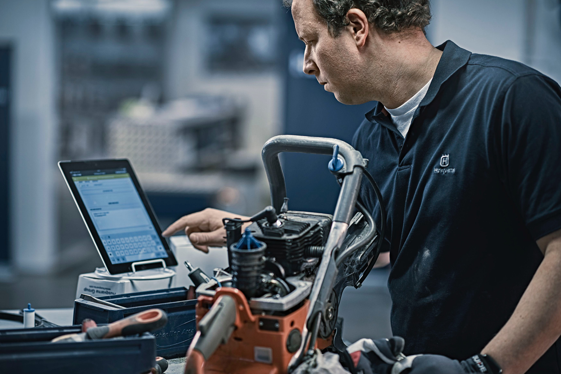 A person in a Husqvarna shirt interacts with a tablet next to engine parts in a workshop.