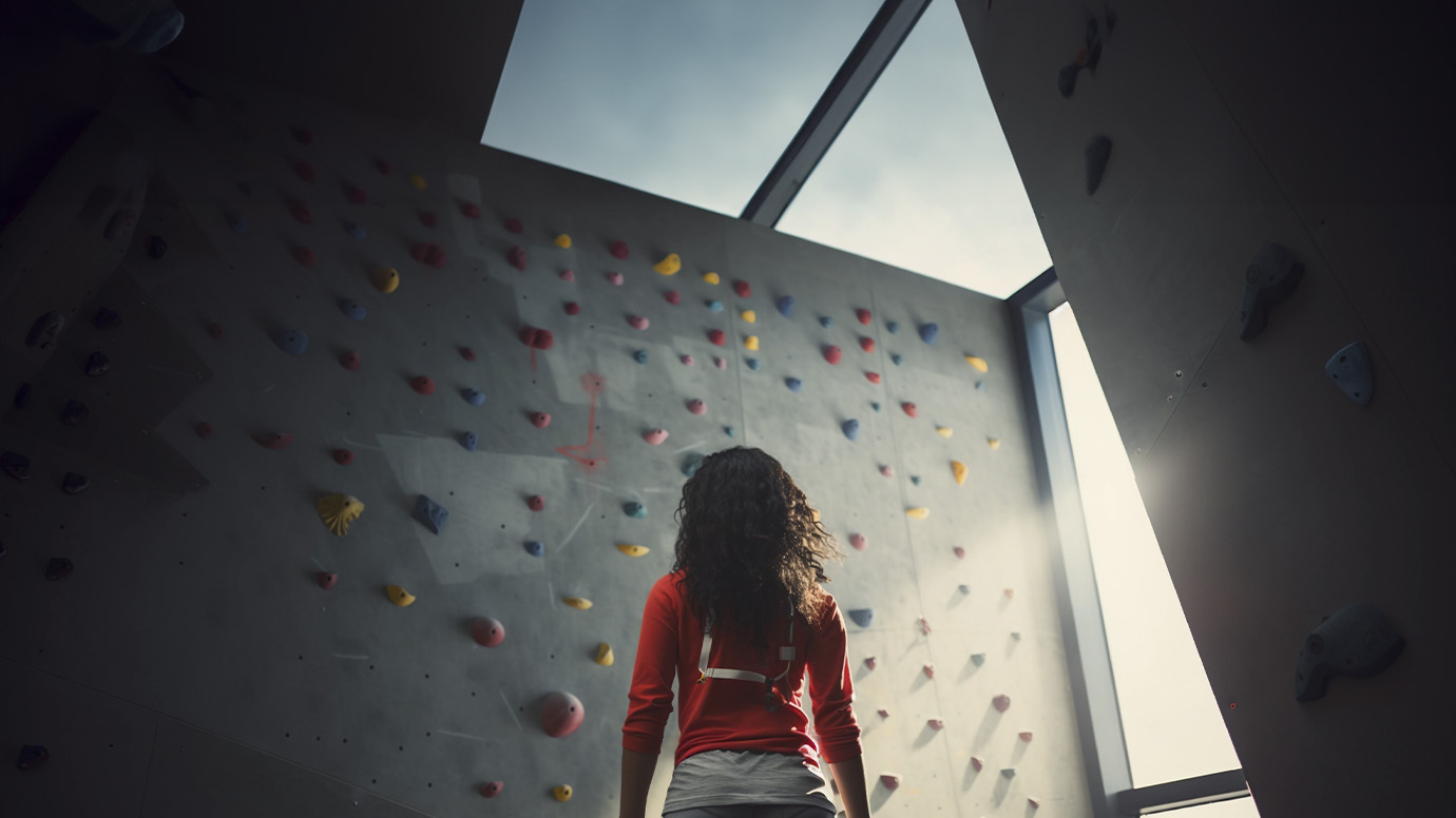 Person in a red shirt facing an indoor climbing wall near a large window, with colorful holds scattered across the wall.