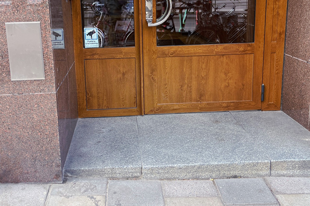 Wooden door with glass panel, surrounded by brown stone walls, with a step-up entrance on a paved walkway.