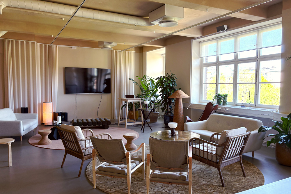 Stylish living room with wicker furniture, large windows, indoor plants, wall-mounted TV, and soft lighting. Neutral color palette.