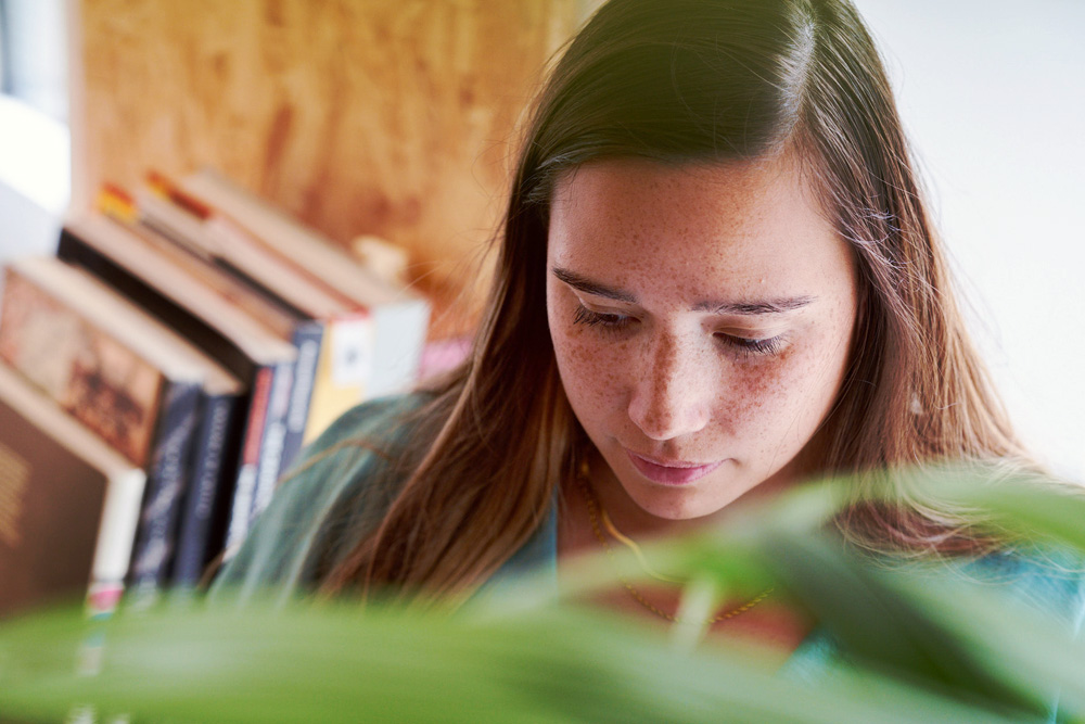Books stacked on a wooden shelf in soft focus, with green leaves in the foreground.