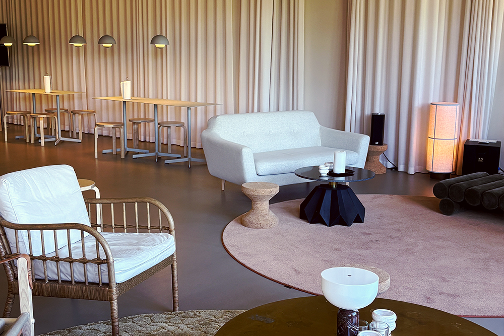Modern lounge area with a white sofa, wicker chair, black table, cork stools, and dining tables in the background. Soft lighting and curtains.
