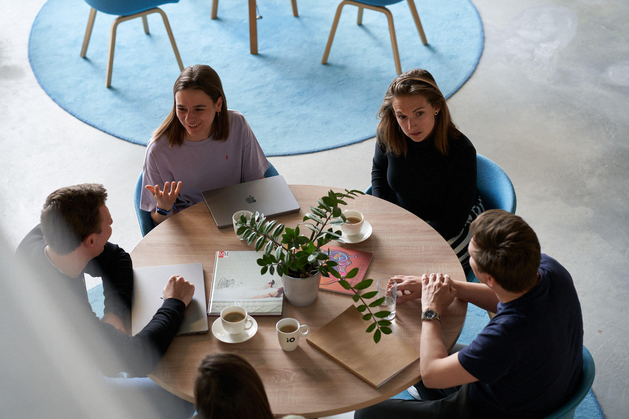 Young adults in a collaborative meeting around a round wooden table with blue chairs. A potted plant, coffee cups, and notebooks sit on the table while a laptop is being used. A blue circular rug is visible in the background.