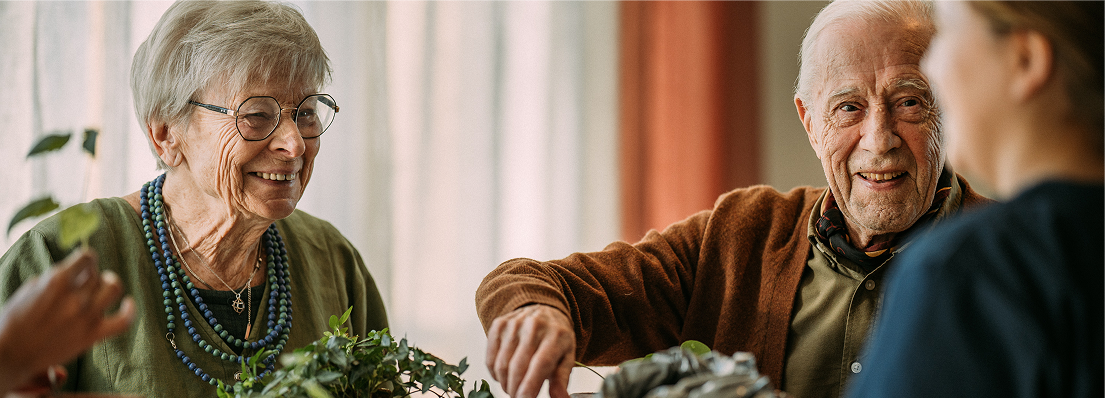 Elderly couple with caregiver smiling warmly together in bright, plant-filled living room