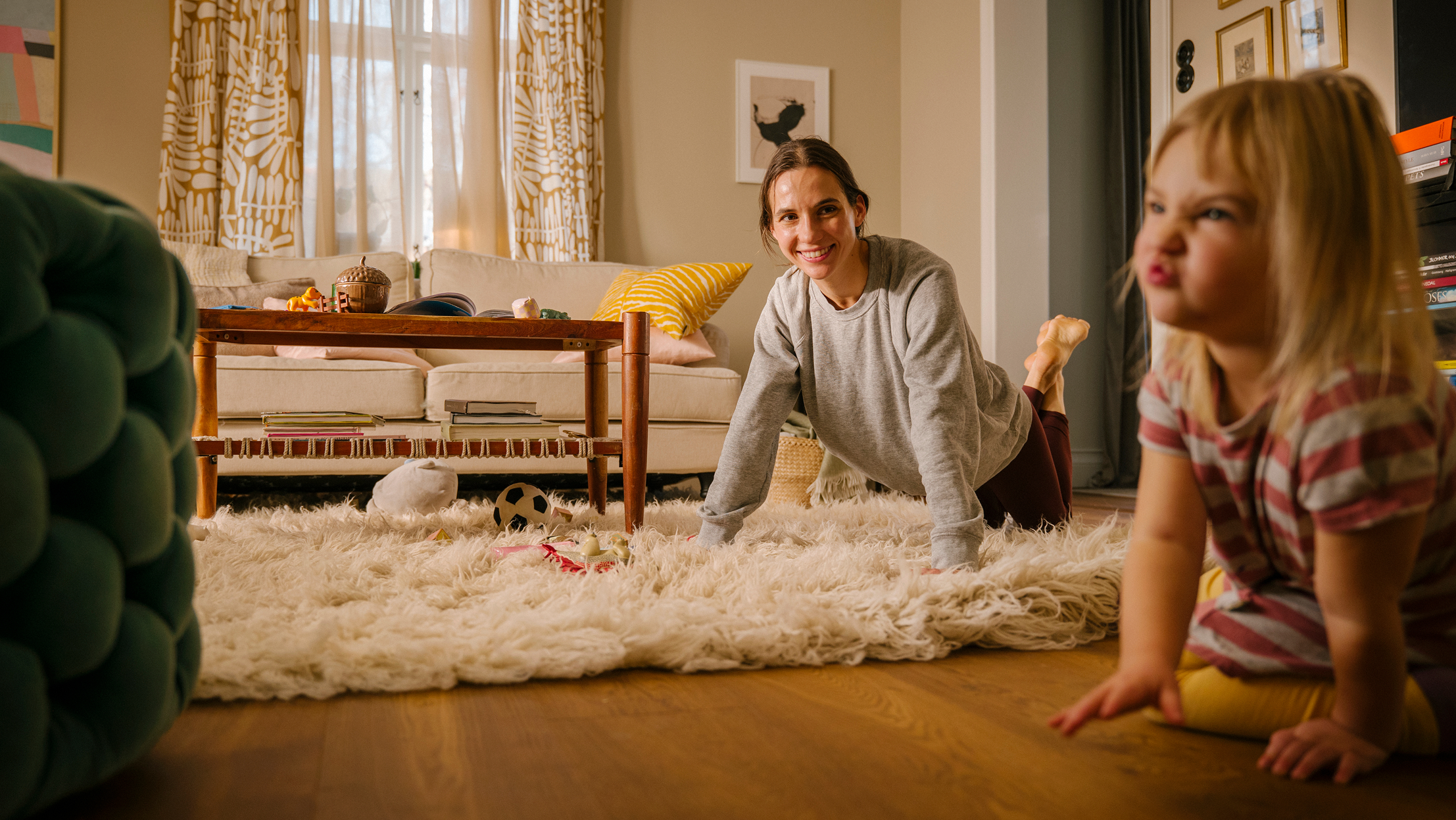 Mother doing push-ups on white fluffy rug in living room, smiling while child plays nearby