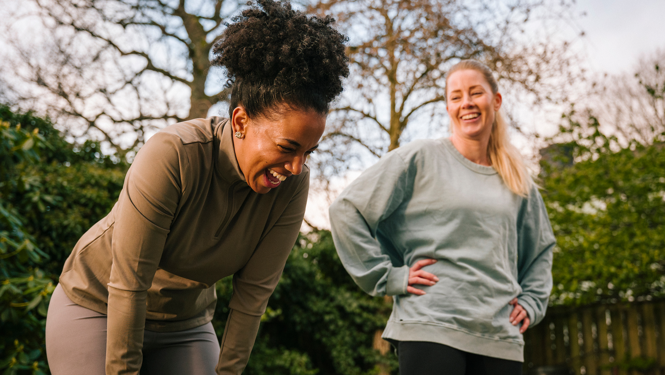Two women laughing during outdoor exercise break in park with trees in background