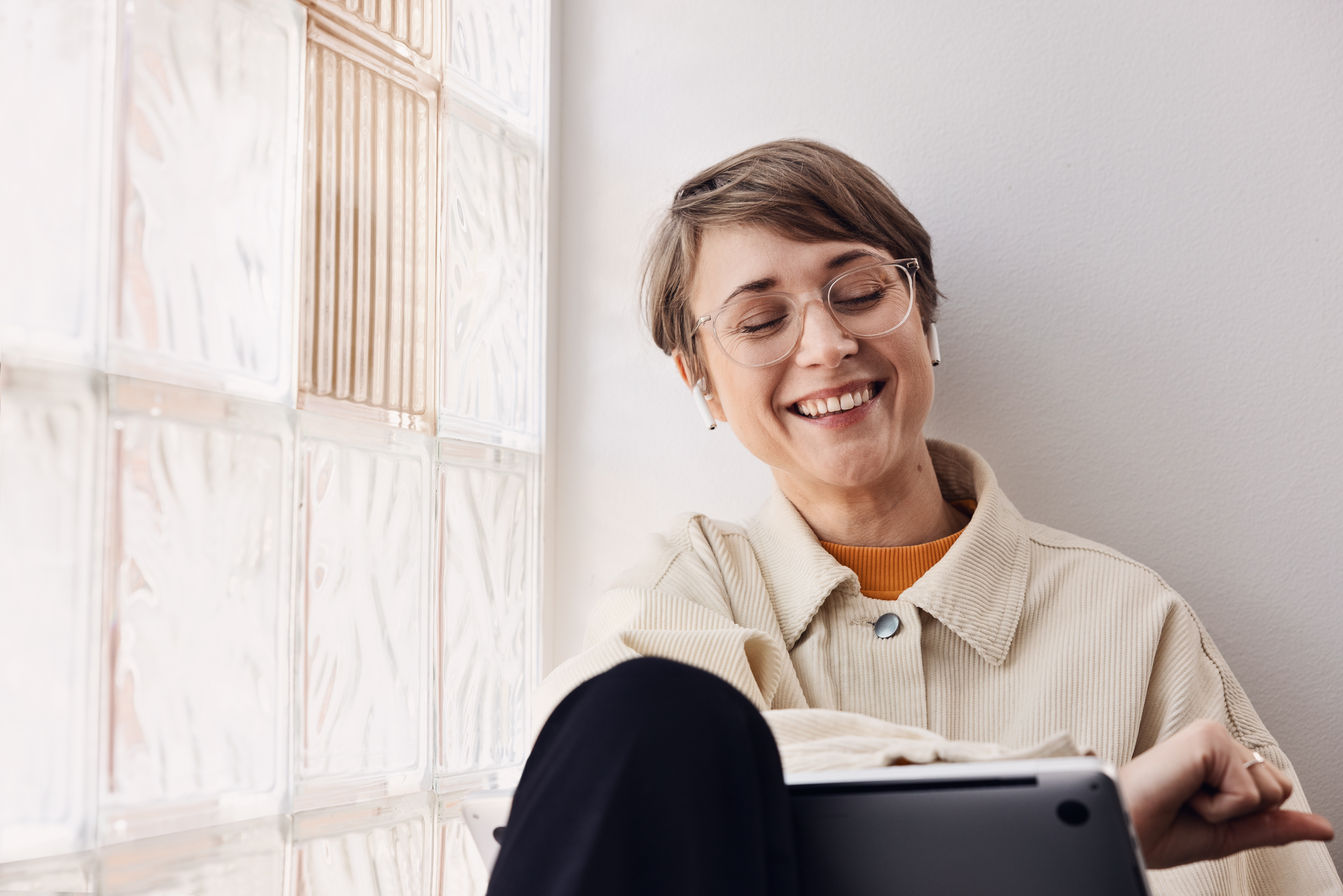Default  A person with short brown hair and round glasses smiles while using a tablet device. They're wearing a cream corduroy jacket with an orange top underneath, sitting beside a textured glass block wall in a bright, minimalist setting.
