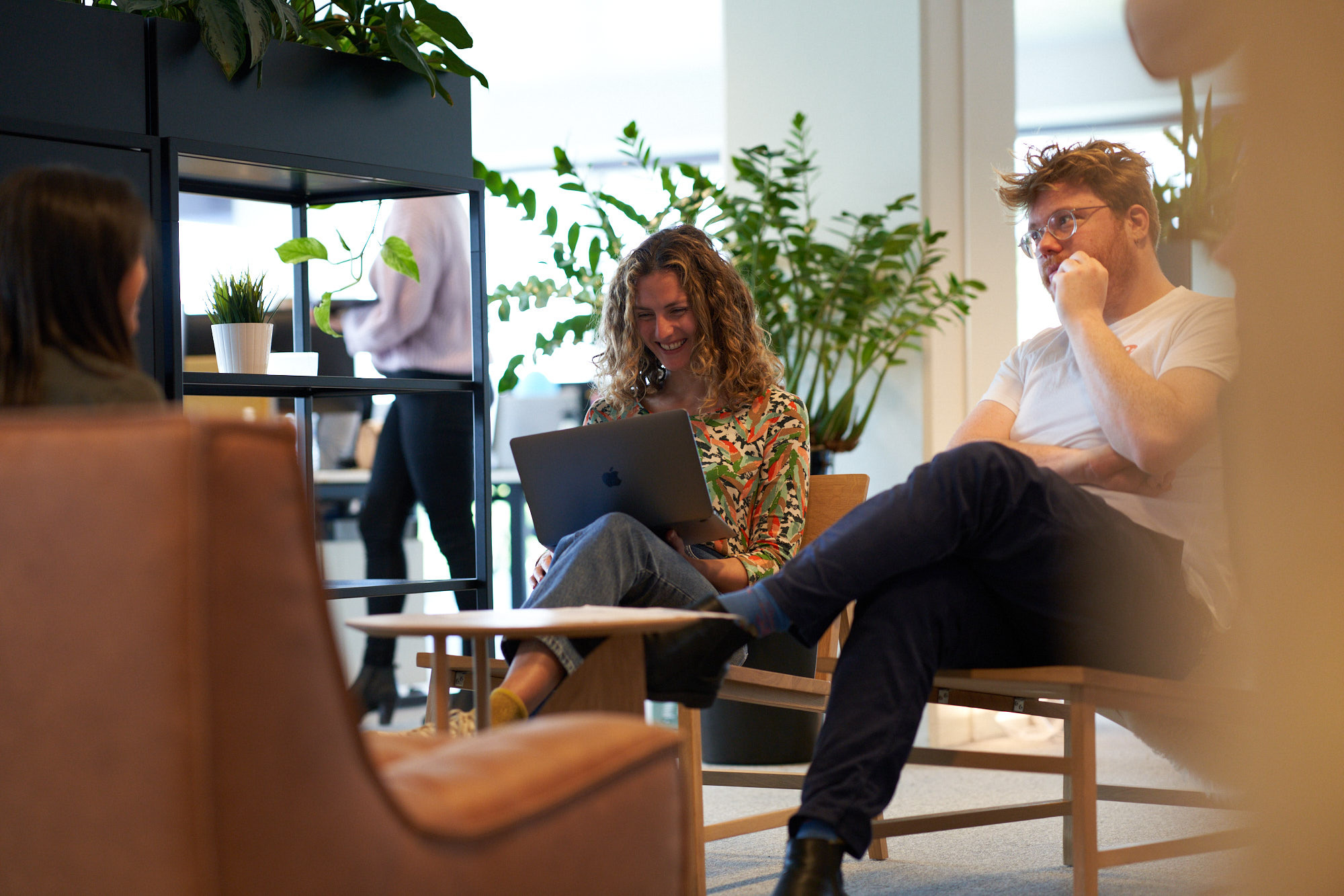 Two people seated in a modern office space, one using a laptop, surrounded by green plants and contemporary furniture.