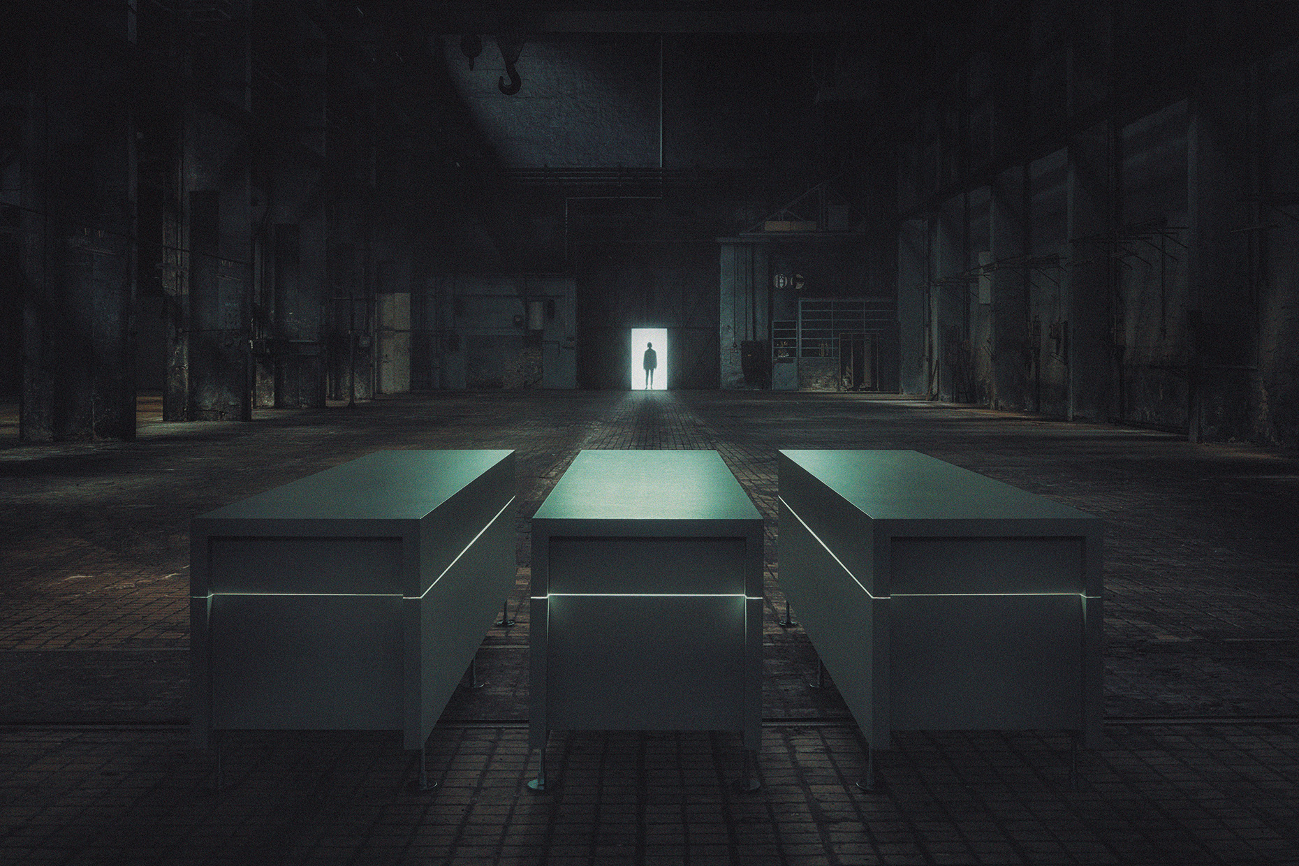 Dimly lit warehouse interior with three illuminated rectangular tables in foreground; silhouette of person in a bright doorway at center.