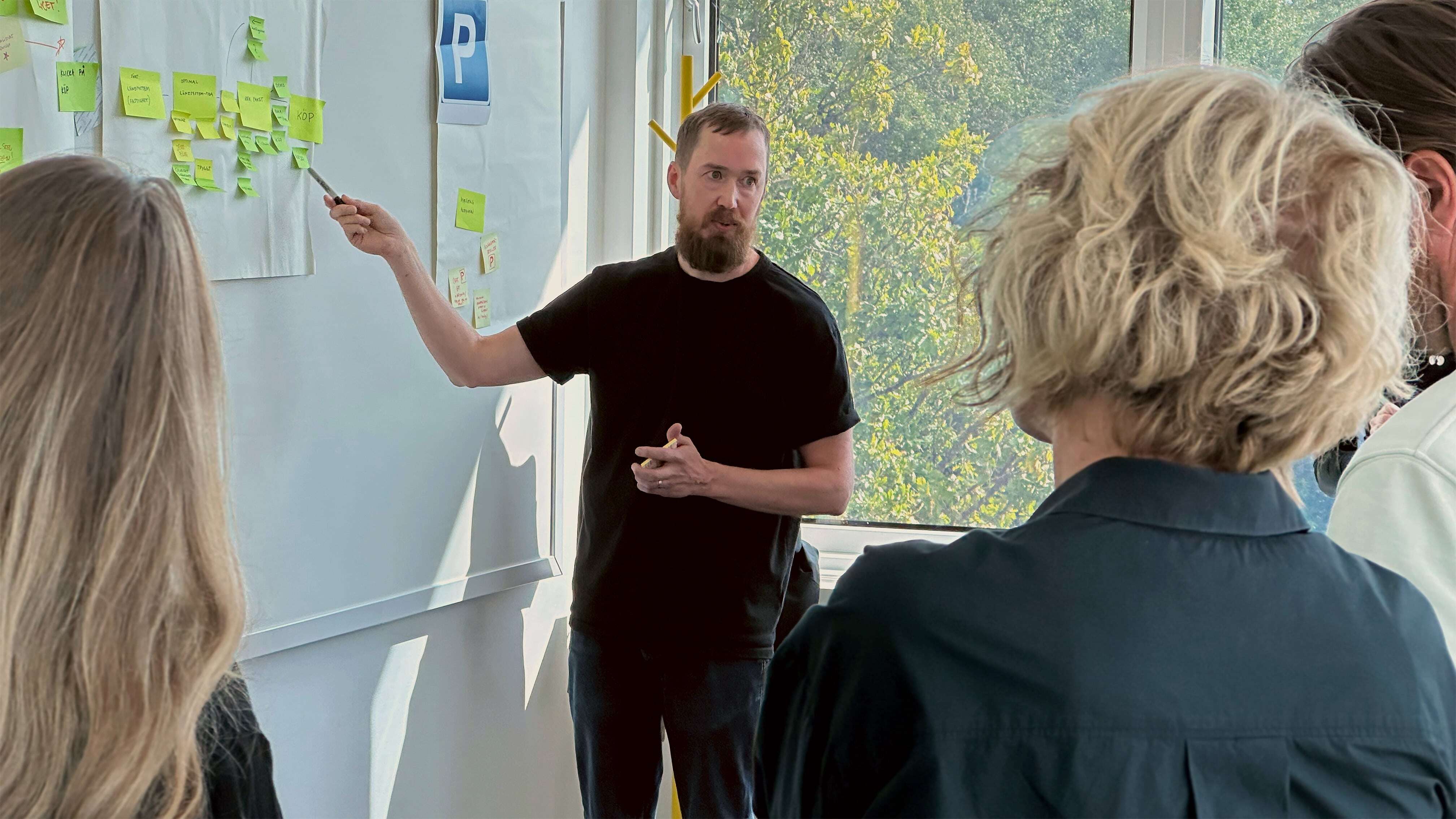 Three people listen to a presentation with sticky notes organized on a whiteboard near a window with trees visible outside.