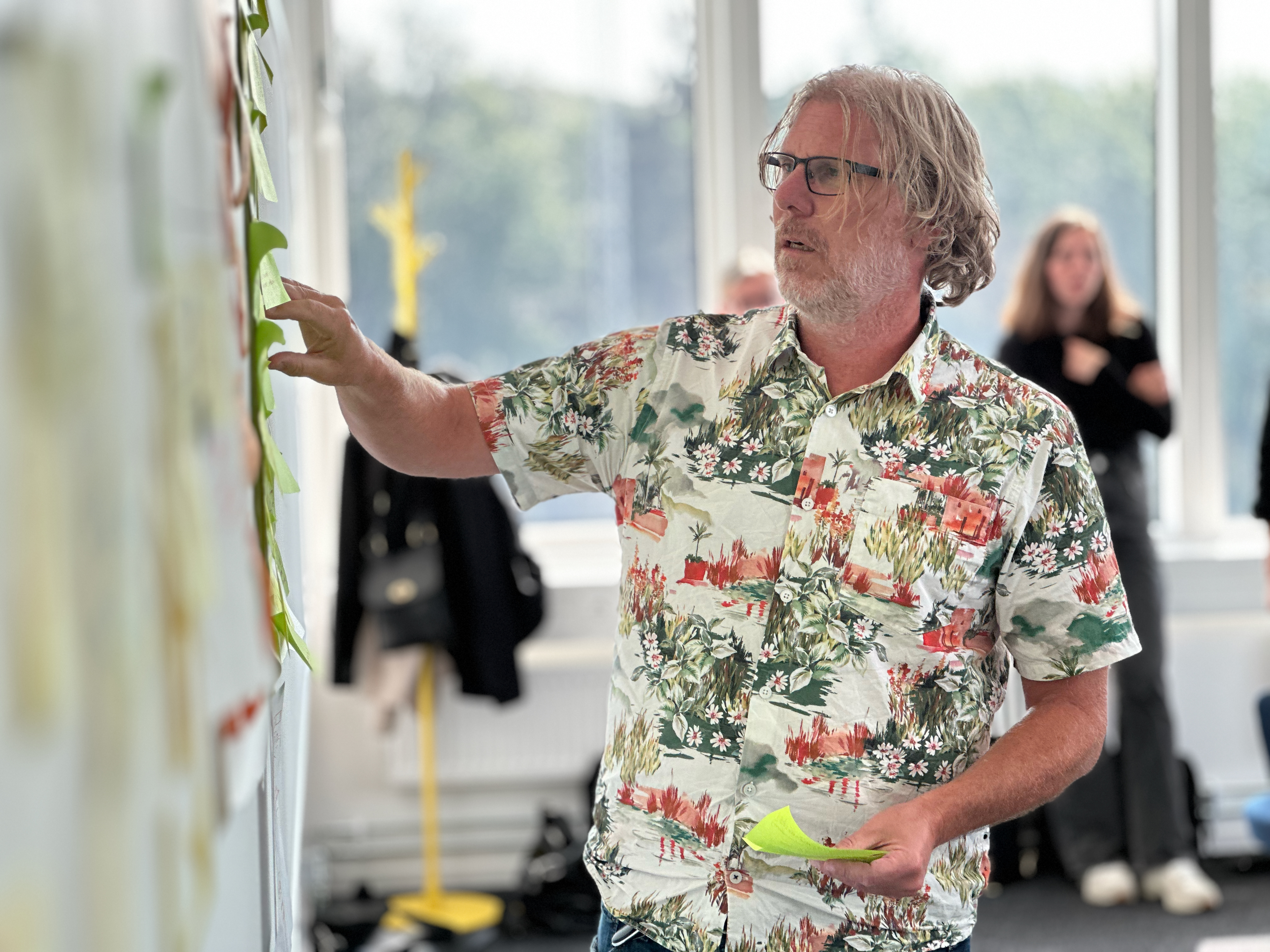 Man in a floral shirt placing a sticky note on a board, with a blurred background featuring coats and a yellow coat rack.