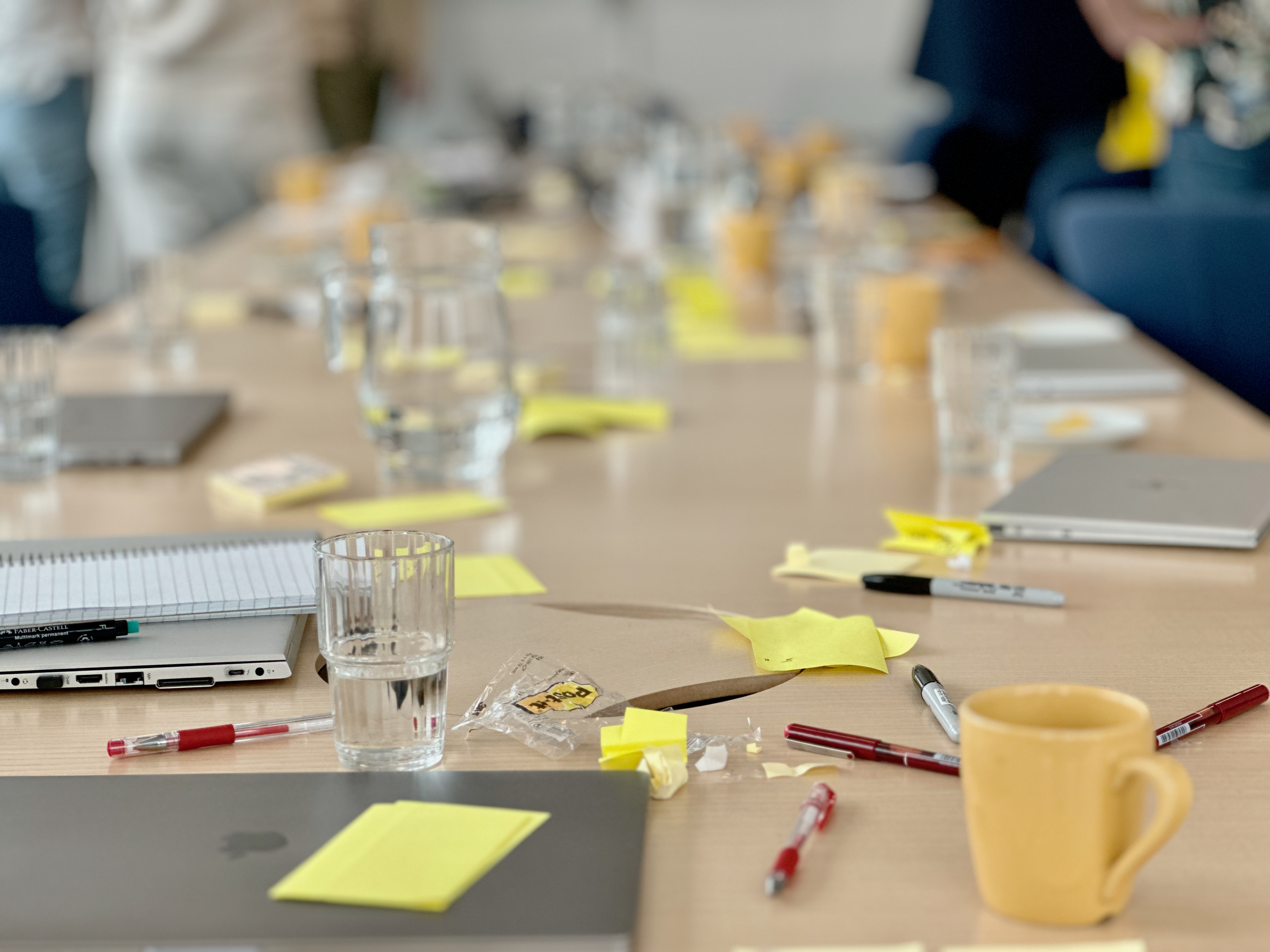 Conference table with laptops, notebooks, pens, water glasses, and scattered yellow sticky notes; people standing in the background.