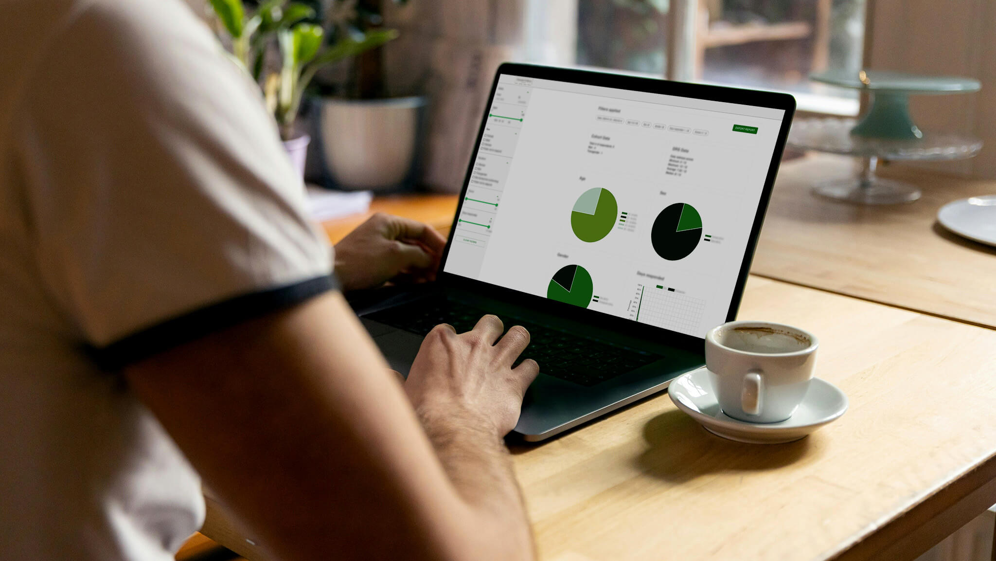 Person typing on a laptop displaying green pie charts while sitting at a wooden table with a cup of coffee nearby.