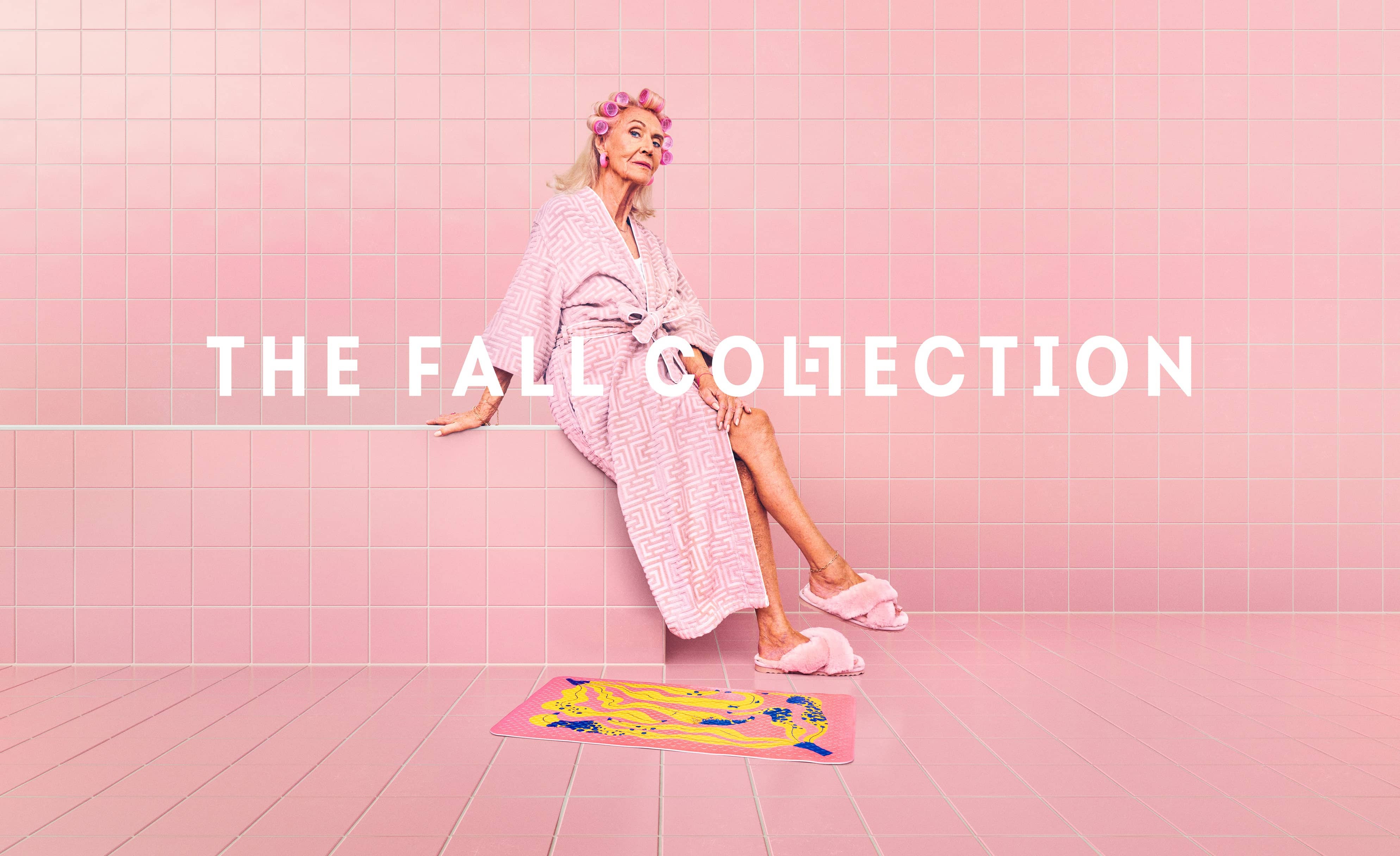 Woman in pink robe and slippers sitting in a pink-tiled room; text reads 'The Fall Collection,' with a colorful mat on the floor.