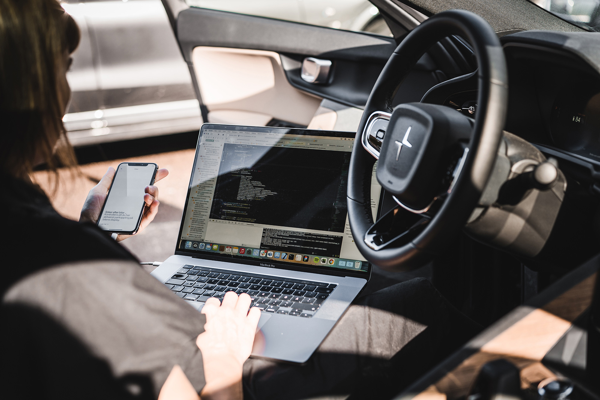 Person in car using a laptop showing code and holding a smartphone, with a focus on programming work and multitasking.