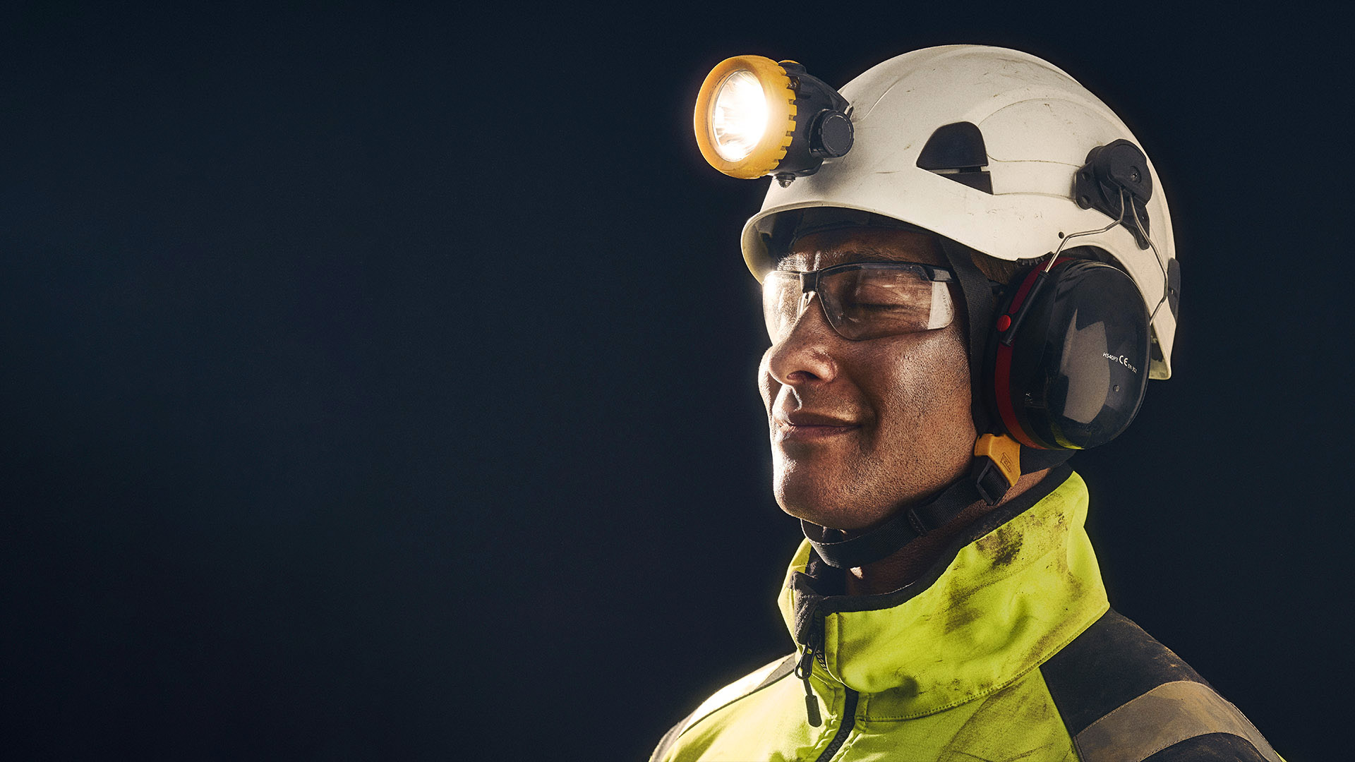 Person wearing a white safety helmet with a headlamp and earmuffs, dressed in a high-visibility jacket.