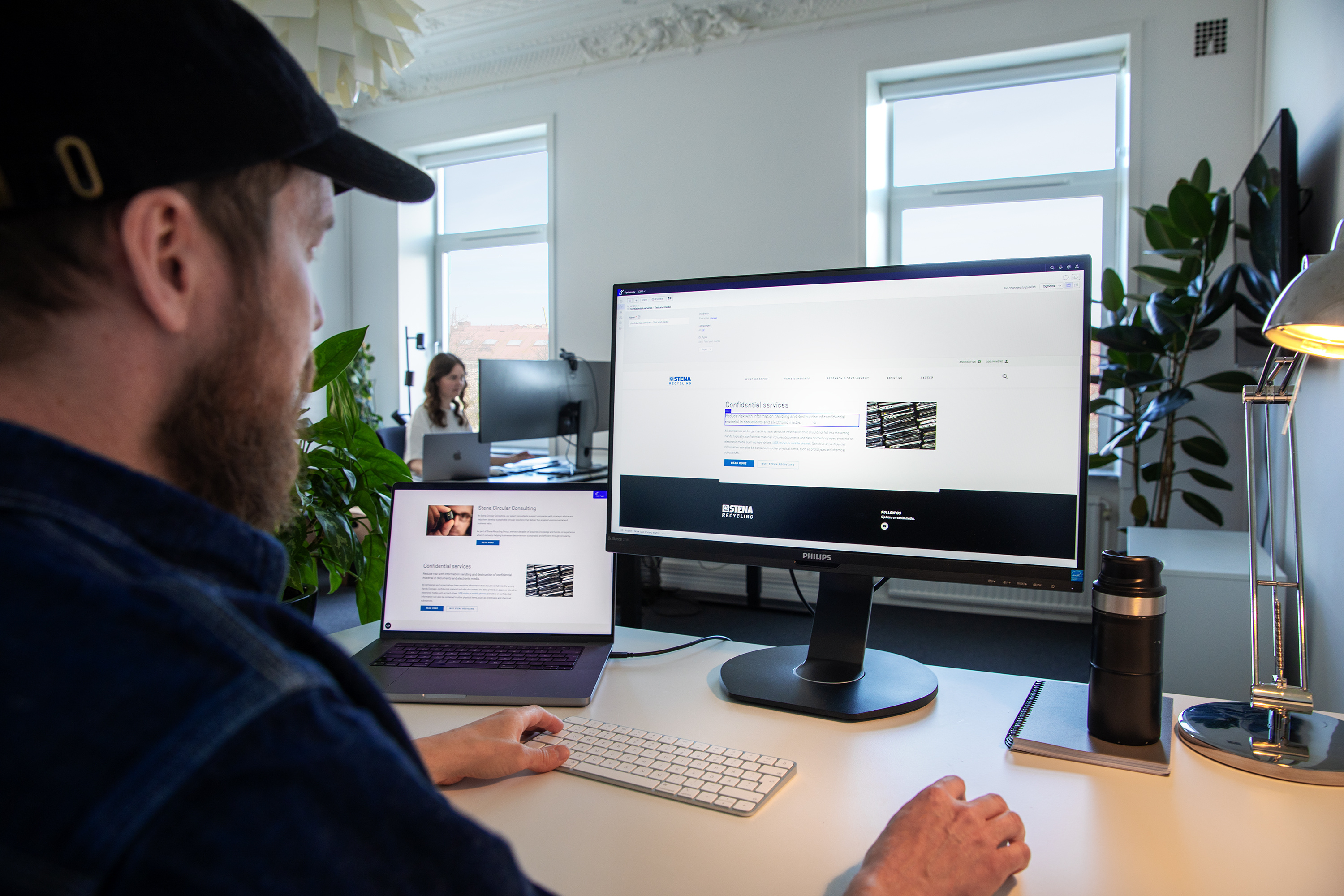 Person working at a desk with a large monitor and laptop displaying website content in a bright office space.