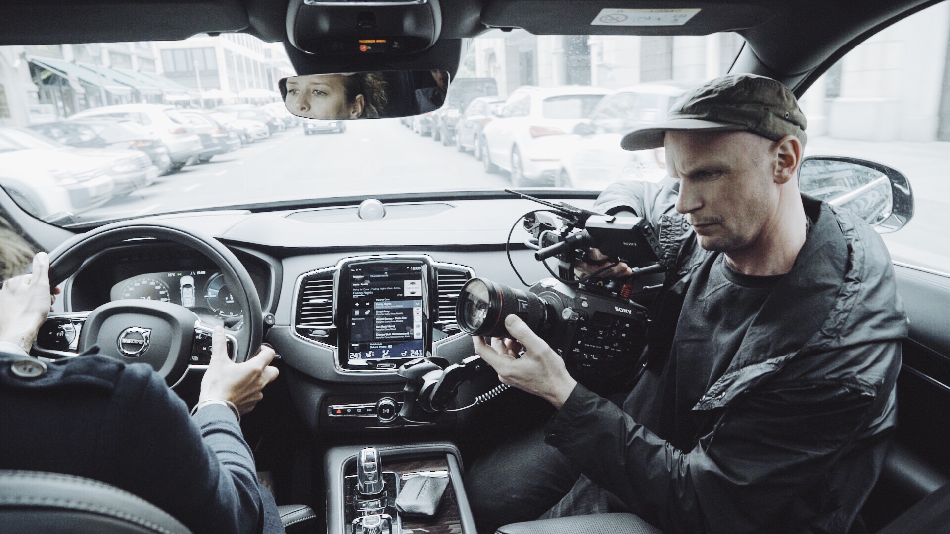 Person driving a Volvo while another films with a Sony camera. Central screen displays navigation and media options.