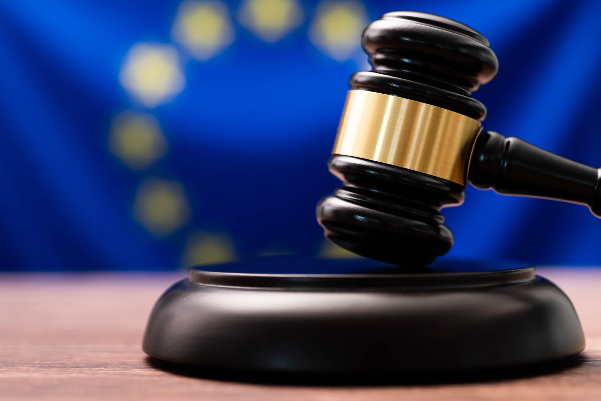 A judge's gavel rests on a wooden surface with the EU flag, blue with yellow stars, blurred in the background.