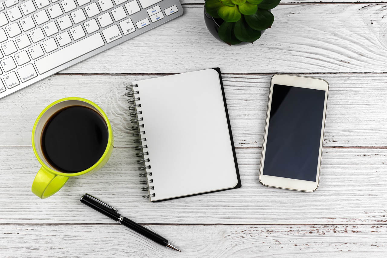 Desk setup with a keyboard, succulent plant, smartphone, spiral notebook, pen, and green mug of black coffee on a wooden surface.